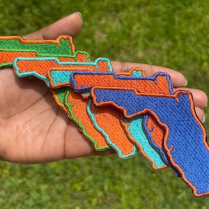 May include: A hand holds a collection of embroidered patches in the shape of the state of Florida. The patches are in various colors, including green, orange, blue, and teal, with orange borders. The patches are likely for clothing or accessories.