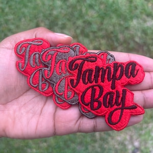 May include: A set of five embroidered patches featuring the words "Tampa Bay" in a red and black script font. The patches are made of a soft, textured fabric and are perfect for adding a touch of team spirit to your belongings.