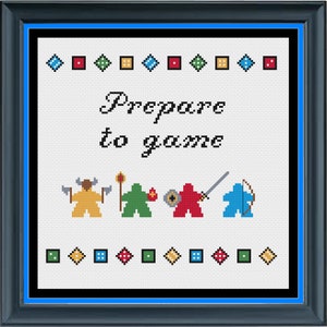 Op de afbeelding: Borduurpatroon met een witte achtergrond en de tekst "Prepare to game" in zwart cursief schrift. Onder de tekst staan vier kleurrijke spelstukken en een rij dobbelstenen boven en onder het patroon.