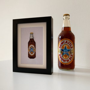 May include: A framed cross-stitch of a brown bottle with a label that says "Newcastle Brown Ale". The bottle is next to a real bottle of Newcastle Brown Ale.