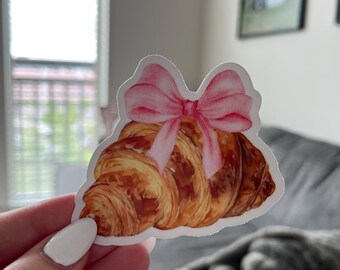 May include: A sticker depicting a watercolour illustration of a croissant adorned with a pink bow. The croissant is a golden brown colour with detailed textures, and the bow is a soft pink. The sticker is held by a person.