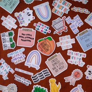 May include: A collection of colorful stickers with various phrases and designs. Some of the stickers feature rainbows, peaches, dogs, and cacti. The stickers are on a wooden surface.