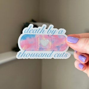 Puede incluir: Una pegatina blanca con un diseño tie-dye rosa y azul que se asemeja a una venda. La pegatina tiene un pequeño corazón blanco en el centro y el texto "death by a thousand cuts" en azul.