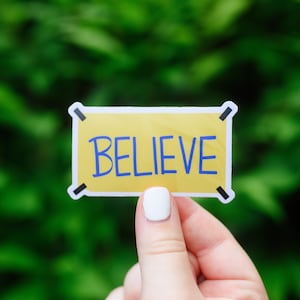 Può includere: Un adesivo giallo con la scritta "BELIEVE" in blu. L'adesivo ha un bordo bianco e dettagli neri agli angoli. Lo sfondo è un verde sfocato.