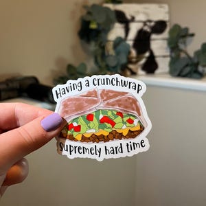 May include: A white sticker with a cartoon drawing of a taco with the text "Having a crunchwrap supremely hard time".