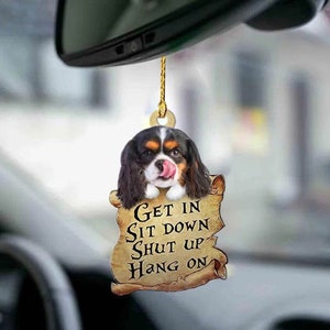 May include: A car air freshener with a Cavalier King Charles Spaniel dog peeking out of a scroll with the text "Get in Sit down Shut up Hang on".