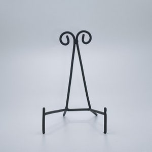 May include: A black metal easel stand with a decorative scroll design at the top. The stand has three legs and is designed to hold a plate or other flat object.