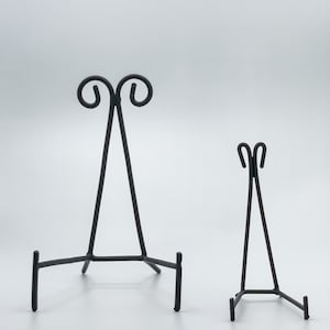 May include: Two black metal display stands for artwork or photos. The larger stand has a decorative scroll design at the top. The smaller stand has a simple design.