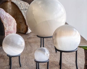 May include: Four selenite spheres of varying sizes on black metal stands. The spheres are translucent white with a slight shimmer.