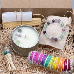 May include: A gift box filled with items for a spiritual practice. The box contains a white candle with crystals, a sage smudge stick, a palo santo stick, a selenite crystal, a green and purple fluorite bracelet, and a small glass bottle of matches.