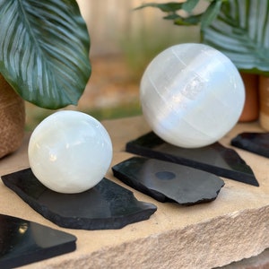 May include: Two white selenite spheres of different sizes on black slate coasters. The larger sphere is on a larger coaster, and the smaller sphere is on a smaller coaster.
