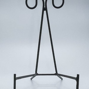 May include: Black metal easel stand with a triangular frame and three legs. The stand is designed to hold a plate or other flat object.