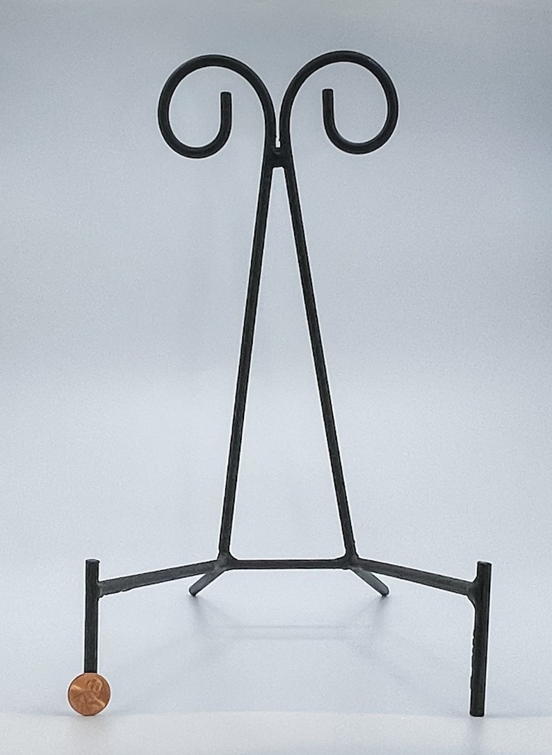 May include: Black metal easel stand with a triangular frame and three legs. The stand is designed to hold a plate or other flat object.