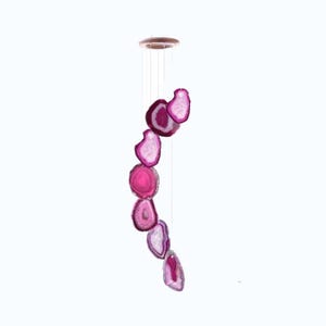 May include: A decorative wind chime featuring slices of pink agate. The agate slices are suspended from a wooden ring by thin strings. The agate slices vary in size and shape, with a mix of pink and white hues. The wind chime is set against a white background.