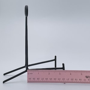 May include: Black metal stand with a curved top and three legs. The stand is approximately 7.6 cm tall and 12.7 cm wide.