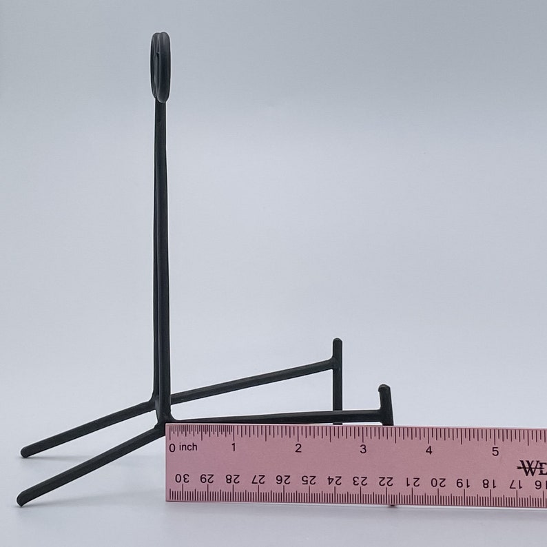 May include: Black metal stand with a curved top and three legs. The stand is approximately 7.6 cm tall and 12.7 cm wide.