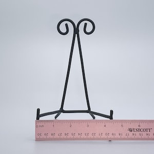 May include: Black metal easel stand with a decorative scroll design at the top. The stand is approximately 15 cm tall.