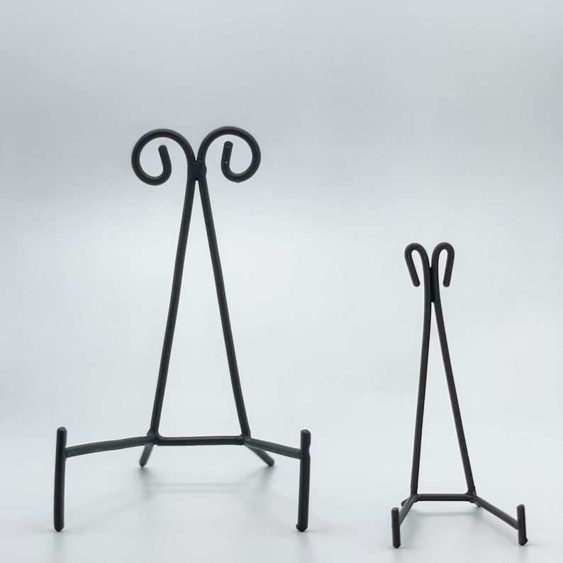 May include: Two black metal display stands for artwork or photos. The larger stand has a decorative scroll design at the top. The smaller stand has a simple design.