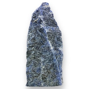 May include: A raw, uncut sodalite gemstone. The stone has a deep blue color with white and gray veining throughout. The rock is roughly rectangular in shape and stands upright against a white background. The surface is rough and textured.