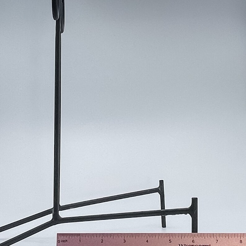 May include: Black metal easel stand with two legs and a curved top. The stand is approximately 18 cm wide.