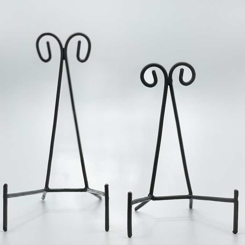 May include: Two black metal display stands with a decorative scroll design at the top. The stands are made of thin metal and have a triangular shape.