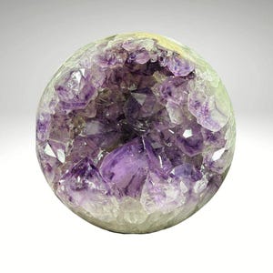 May include: A spherical amethyst geode with a hollow center filled with vibrant purple crystal formations. The outer shell is a pale green color, and the interior crystals are a deep violet. The geode is a natural, decorative item.