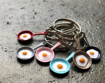 May include: A set of six colorful miniature frying pans with a fried egg design. Each pan is attached to a silver key ring. The key rings are connected to a larger silver ring.
