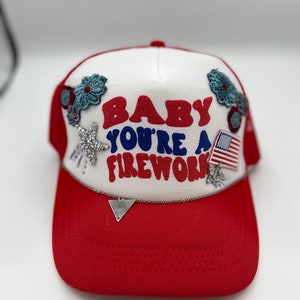 Puede incluir: Gorra de camionero roja y blanca con el texto "BABY YOU'RE A FIREWORK" en rojo y azul. La gorra presenta una visera roja, un panel frontal blanco y una parte trasera de malla roja. Los adornos incluyen una pequeña bandera estadounidense, pedrería en forma de estrella y diseños florales.