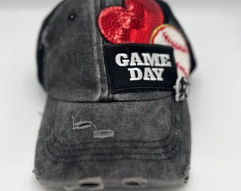 May include: Distressed black baseball cap with a patch that says "GAME DAY". The patch features a red sequined heart and a baseball. The cap has a pre-worn look with distressed details.