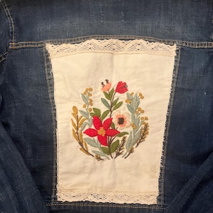 May include: A dark blue denim jacket with a floral embroidered patch on the back. The patch features red, pink, and white flowers with green leaves and gold accents, framed by a cream-coloured lace trim.
