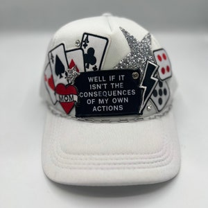 May include: White baseball cap with playing card and dice embellishments. The cap features a black patch with the text "WELL IF IT ISN'T THE CONSEQUENCES OF MY OWN ACTIONS". A silver glitter star and rhinestones add sparkle to the design.