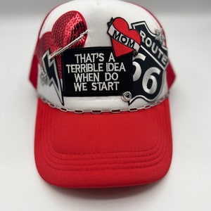 May include: Red and white trucker hat with a red brim. The hat features patches including a sequined heart, a heart with "MOM" on a banner, and a Route 66 sign. The text "THAT'S A TERRIBLE IDEA WHEN DO WE START" is also on the hat.