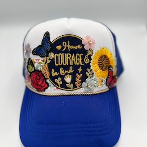 Op de afbeelding: Blauwe en witte truckerpet met bloemen- en vlinderversieringen. De pet heeft de tekst "Have Courage & be kind" in een cirkelvormig ontwerp. De klep is blauw en de kroon is wit.