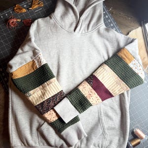 Puede incluir: Una sudadera con capucha gris con mangas de patchwork. Las mangas presentan rayas horizontales de telas doradas, verdes, florales y burdeos. La sudadera tiene un bolsillo de canguro y una capucha con cordón. El fondo incluye una esterilla de corte y carretes de hilo.