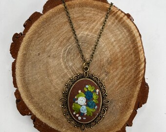 May include: Oval pendant necklace with a bronze-colored chain. The pendant features an embroidered floral design in shades of blue, white, and green on a brown background, set within a decorative bronze frame. The necklace is displayed on a wooden slice.