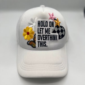 May include: White trucker cap with a curved peak and mesh back. The cap has embroidered patches of a yellow flower, butterfly, star, and a checkered flag. The main patch reads "HOLD ON LET ME OVERTHINK THIS."