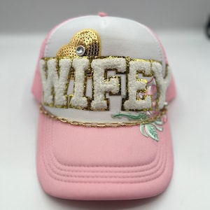 May include: Pink and white baseball cap with the word "WIFEY" in white, fluffy letters. The cap features a gold sequined heart and a floral design. A gold chain accents the brim.