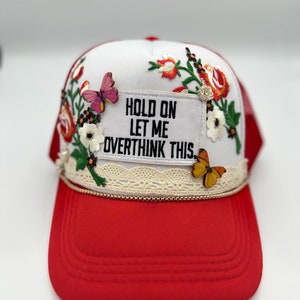 May include: A red and white trucker hat with floral embroidery and butterfly accents. The hat features the text "HOLD ON LET ME OVERTHINK THIS." A cream-coloured lace trim and gold rope detail the brim.
