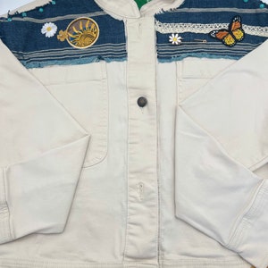 May include: Cream-coloured jacket with denim accents and decorative patches. The jacket features a round sun patch, a butterfly patch, and daisy patches. The denim sections are embellished with turquoise beads and lace trim. The bottom hem is trimmed with a mustard-coloured lace.