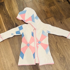 Op de afbeelding: Een witte gewatteerde jas met capuchon, met een geometrisch patroon van roze en blauwe vormen. De jas heeft een roze bies en knopen aan de voorkant. De mouwen zijn lang. Ligt op een houten oppervlak.