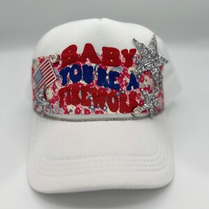 Könnte beinhalten: Weisse Trucker-Mütze mit gebogenem Schirm. Der Hut trägt den Text "BABY YOU'RE A FIREWORK" in Rot und Blau, mit einem Blumenmuster und einer amerikanischen Flagge. Verziert mit silbernen Glitzersternen und Strasssteinen.