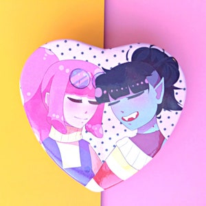 May include: Heart-shaped pin featuring two cartoon characters. The character on the left has pink hair, wears glasses, and a blue and purple sweater. The character on the right has blue hair, wears a white sweater, and has fangs. The background is white with black polka dots.