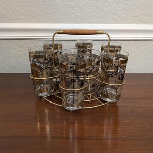 May include: A set of six vintage highball glasses with a gold and black map design, held in a gold-toned metal carrier with a wooden handle. The glasses have an "N" logo.
