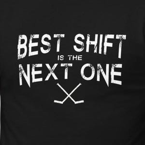 May include: Black t-shirt with the white text "BEST SHIFT IS THE NEXT ONE" and two crossed hockey sticks. The text has a distressed, textured appearance. The shirt is short-sleeved and appears to be made of a soft material.