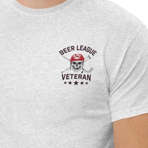 Puede incluir: Camiseta gris claro con un pequeño gráfico en la parte superior izquierda del pecho. El gráfico incluye una calavera con un casco de hockey, palos de hockey cruzados y las palabras "BEER LEAGUE VETERAN" con cinco estrellas debajo. La camiseta tiene mangas cortas.