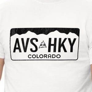 May include: White t-shirt featuring a black rectangular design with the text "AVS HKY" and "COLORADO". The design includes a mountain range graphic and a triangle. The shirt is made of a soft, comfortable material.