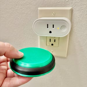 May include: A round green and black button remote held in front of a white wall outlet. Above the outlet is a white smart plug with a single power button and a small yellow indicator light, plugged into the top socket of the wall plate.
