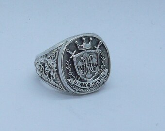 May include: A silver ring with a detailed design featuring a shield with a crown, swords, and the text "UT AMOR AMENTUR".