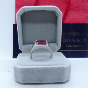 May include: A silver ring with a red gemstone in a gray velvet ring box. The box has a blue lid with the text "Certificato di Garanzia".