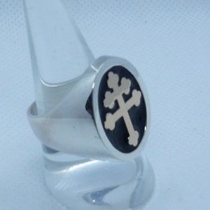 May include: A silver ring with a black enamel inlay featuring a gold-toned cross with four points. The cross is a traditional symbol of faith.